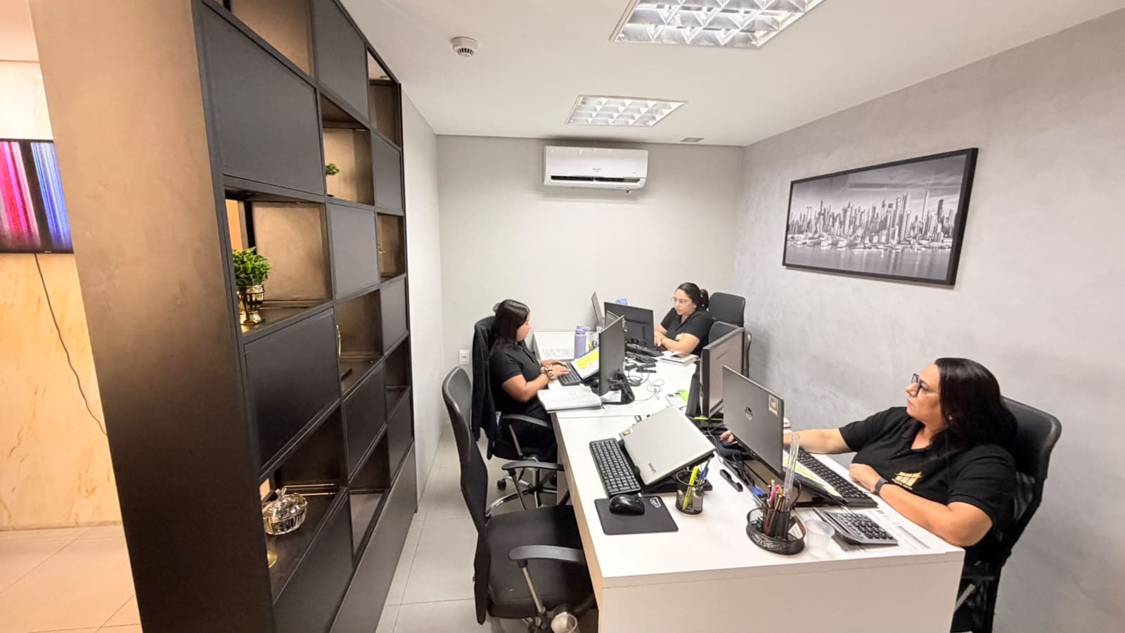 Três integrantes da equipe trabalhando em seus computadores, ao lado de uma estante escura