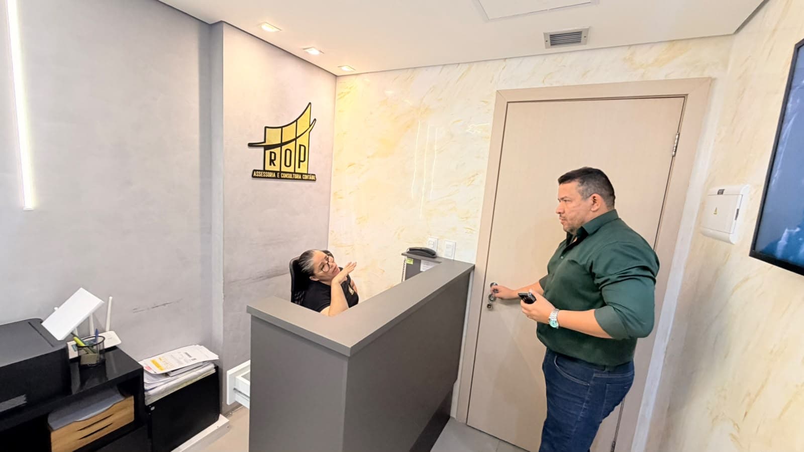 Recepção da ROP, com o logotipo dourado na parede, a recepcionista no balcão e o sócio à porta