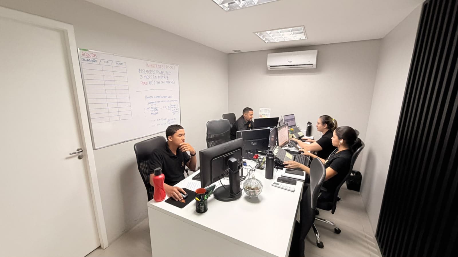 Equipe à mesa de trabalho, com um quadro branco de agenda de declarações e prazos fiscais na parede