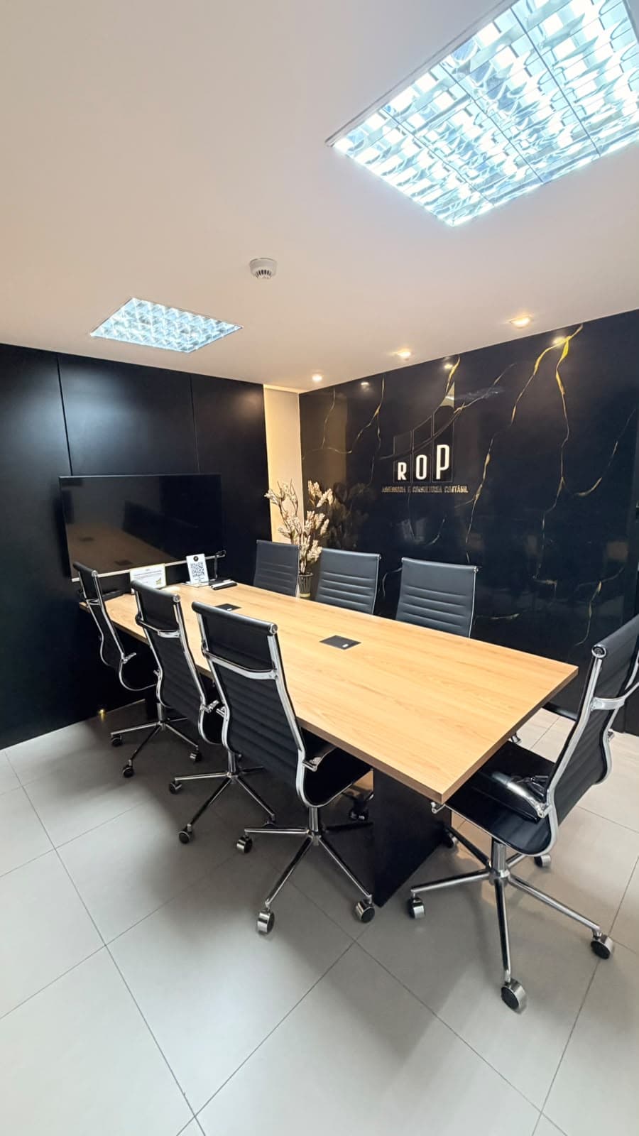Sala de reunião com mesa de madeira, cadeiras pretas e parede de mármore escuro com o logotipo da ROP